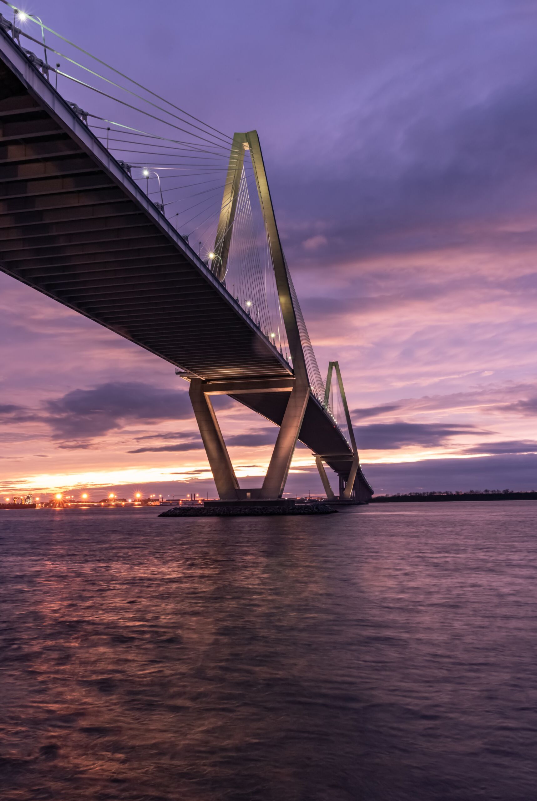 Vertical picture of a bridge on the sea surrounded by lights under a cloudy sky during the sunset